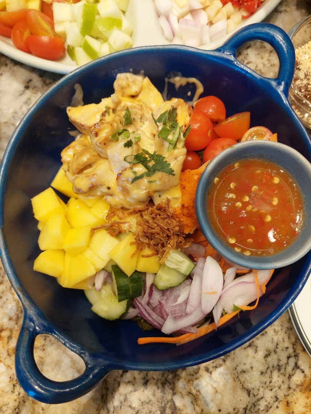 A cozy blue bowl filled with mango, vegetables, chicken, and dipping sauce on a marble table.