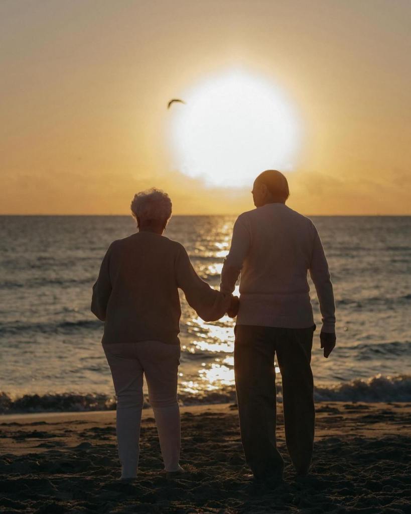 An elderly couple holding hands by the sea at sunset, representing lifelong love and companionship