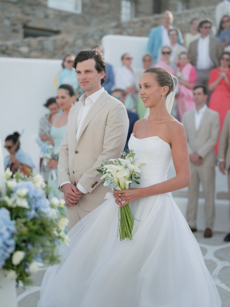 Bride and groom standing together during a wedding ceremony in Santorini, Greece, surrounded by guests and coastal scenery