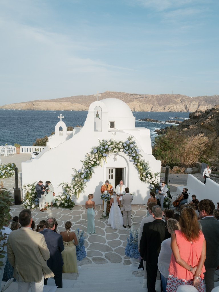 Outdoor wedding ceremony at a white church in Santorini, Greece, overlooking the sea and surrounded by guests