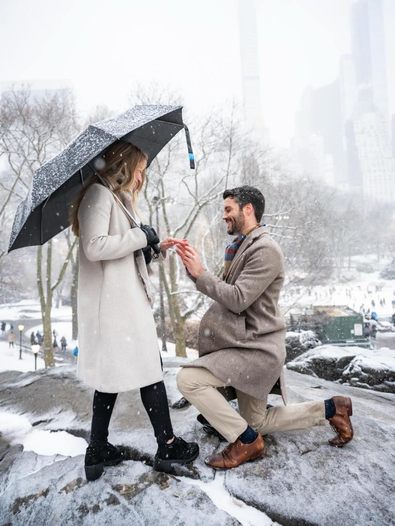 A man kneeling to propose in a snowy park while holding a woman’s hand under an umbrella, capturing a romantic winter proposal moment
